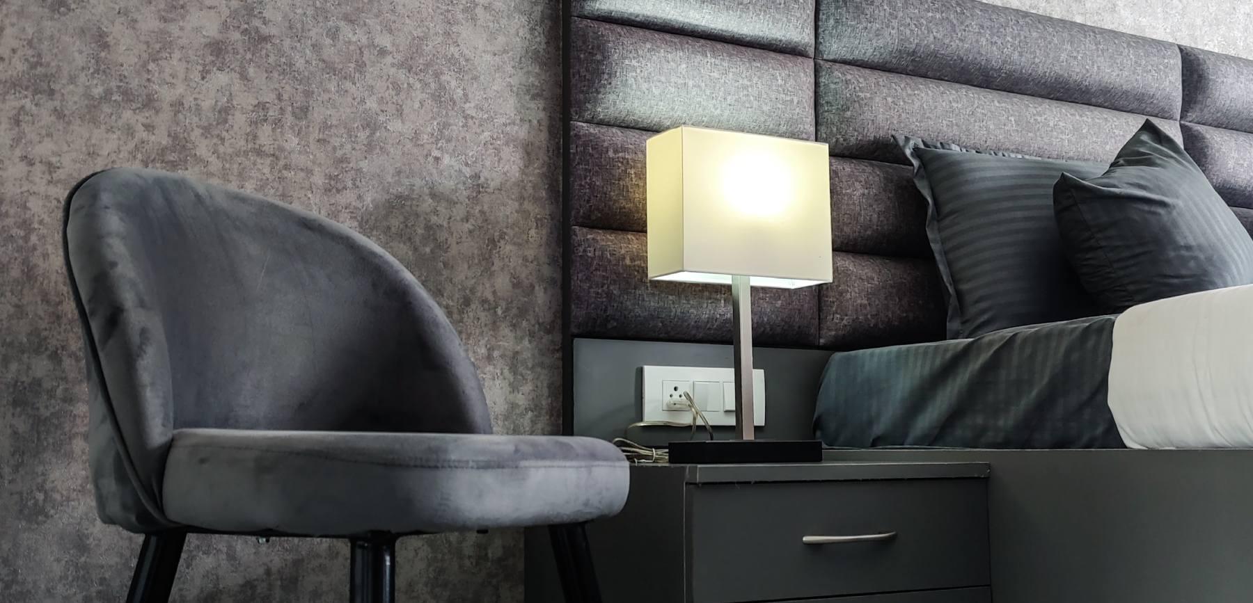 Stylish modern room for rent interior with gray velvet chair, textured wall, and bedside lamp.
