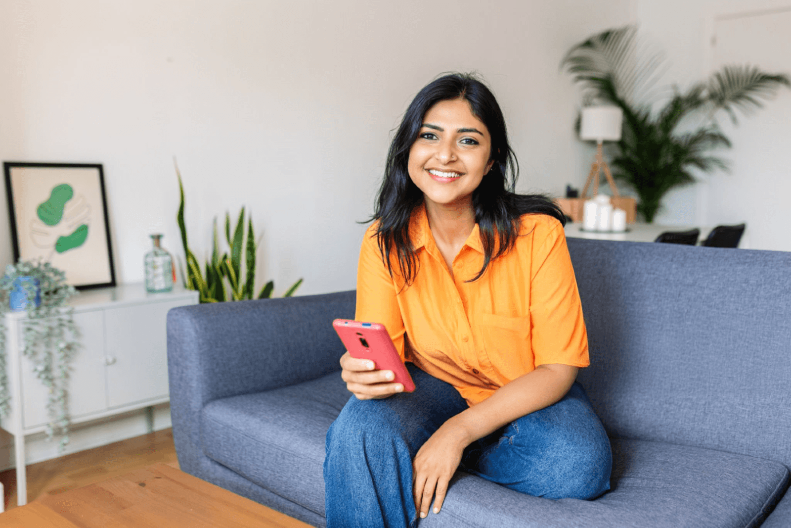 Smiling young woman sitting in a modern shared apartment, using her phone – representing affordable coliving spaces in Hyderabad.