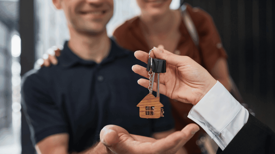 Real estate agent handing over house keys with a house-shaped keychain to a happy couple, representing a successful deal for a 1BHK for rent in HSR Layout.